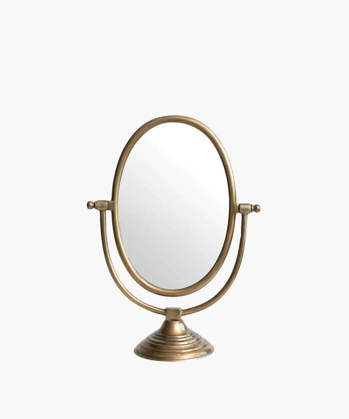 A vintage-style tabletop mirror with an oval shape and bronze finish. It features an adjustable frame resting on a decorative, circular base. | Wildwood Home Co.