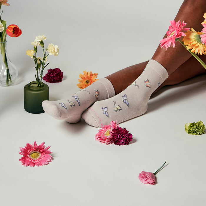 Beige half cushion socks with butterfly designs, surrounded by colorful flowers on a white surface.