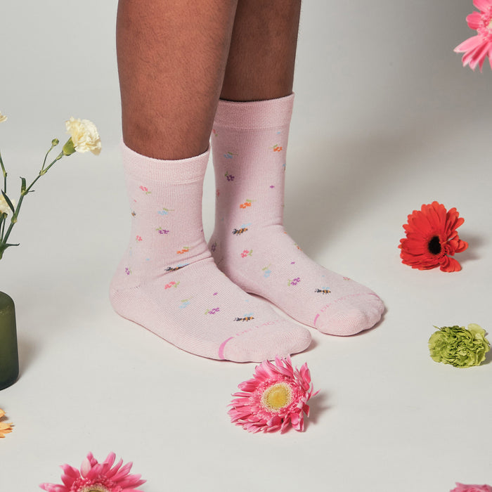 Pink half cushion socks with floral and bee patterns, worn on feet surrounded by flowers.