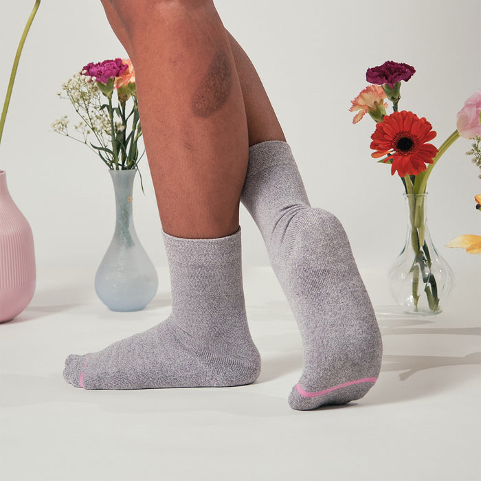 Gray half cushion socks with a pink accent, worn on a model's feet, surrounded by vases of flowers.