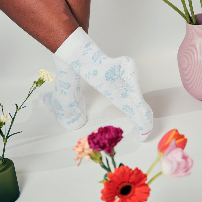 White socks with blue floral patterns, worn on a person's feet, surrounded by colorful flowers and a vase.