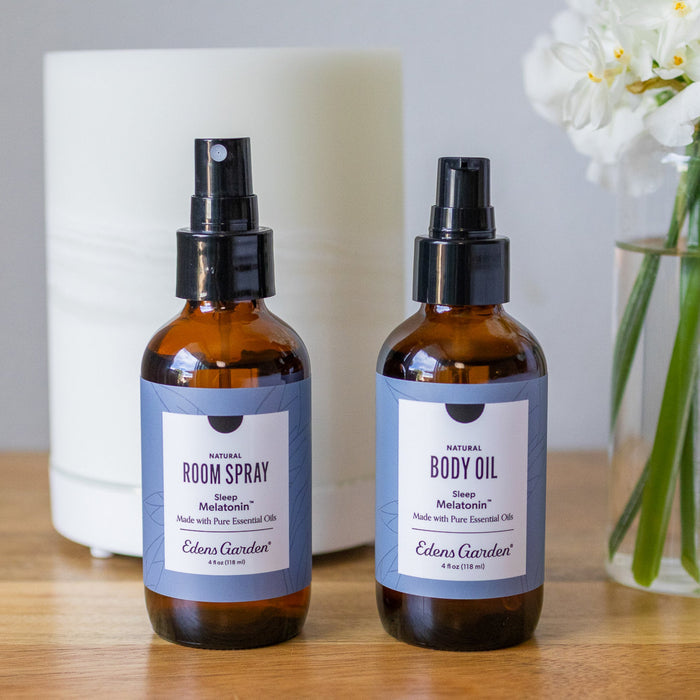 Two bottles of Edens Garden room spray and body oil on a wooden surface with a white candle and flowers in the background.