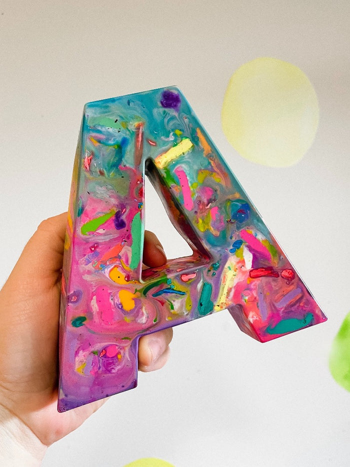 Huge letter A. Image is of an adult hand holding up a Crayon letter A on a white background. Crayon colors are in a mix of pastel pinks and teals. This oversized crayon is in the shape of a large letter A.  This rainbow crayon from crayon shop, Art 2 the Extreme, has been created with pastel colored recycled crayons. This unique gift for kids is larger than an adult hand and perfect for back to school gift ideas or stocking stuffers for kids. Pair with a kids paper pad!