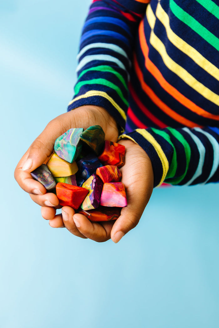 Child holding a handful of multicolored  rock crayons in different hues. Handful has about 15 rainbow rock crayons on a teal blue background.