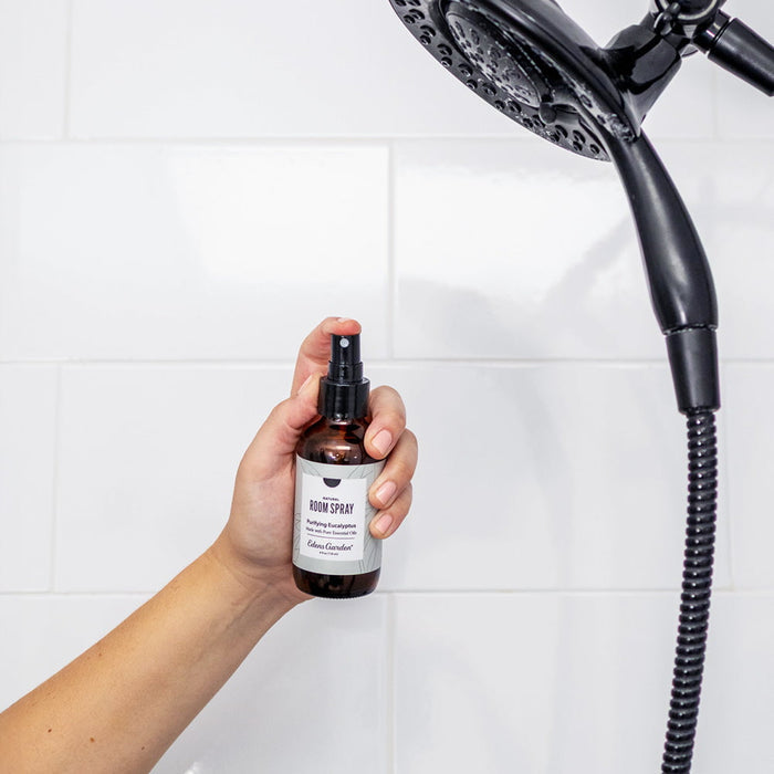 Hand holding a bottle of room spray next to a shower head against a tiled wall.