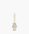A tall white candle in a rounded, beige travertine stone holder with a textured surface. The minimalist design evokes a calm and serene atmosphere. | SW Home