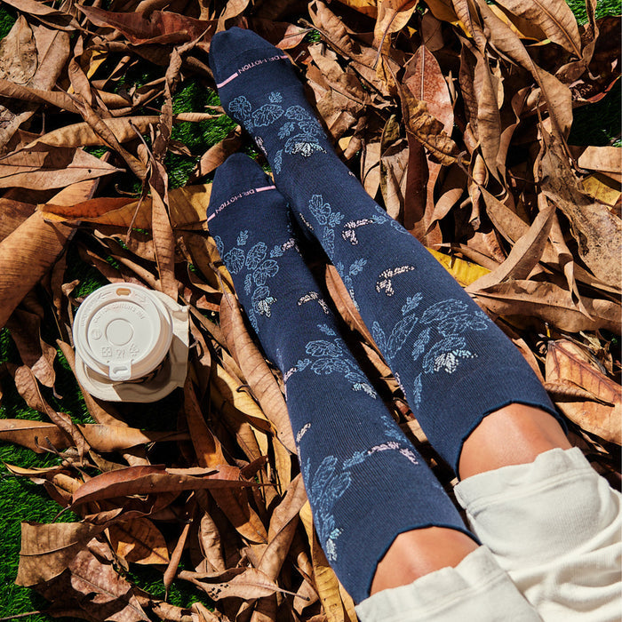 Knee-high compression socks with a navy background and whimsical butterfly and floral designs, resting on dry leaves.